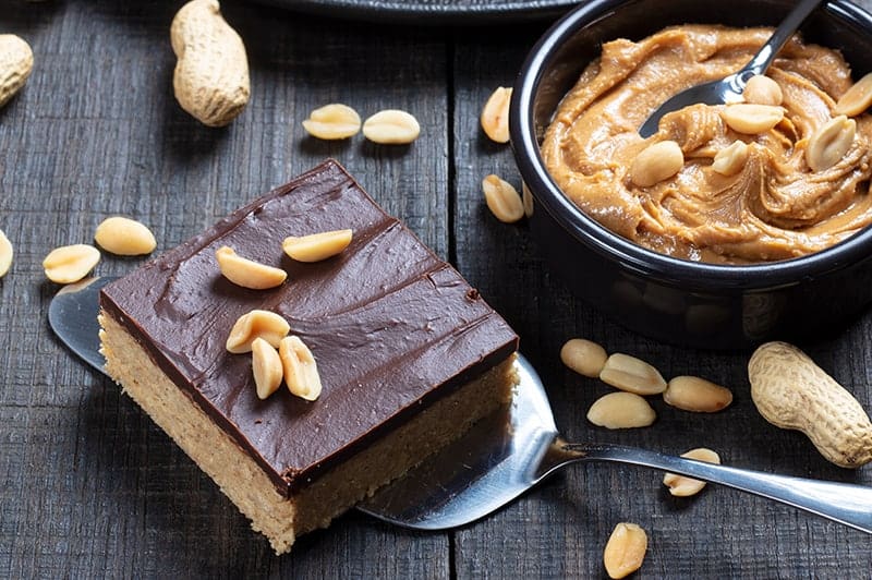 a piece of Peanut Butter Bar in a cake spatula sprinkled with roasted peanut, peanut butter spread on the side