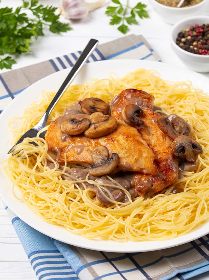 checkered blue kitchen towel underneath white plate with Chicken Marsala on angel hair pasta and a fork on it, parsley leaves and some ingredients on its background