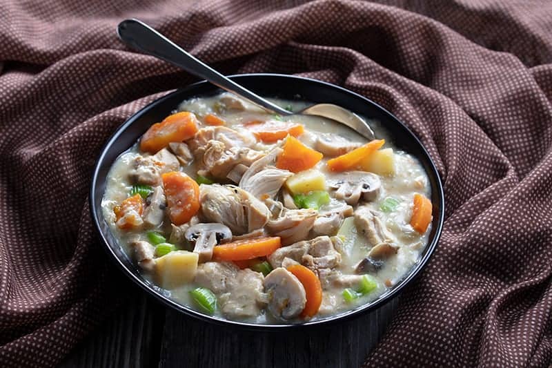 slow cooker chicken stew in a large soup bowl with spoon and a brown tablecloth around it