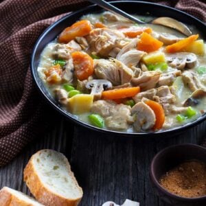 some french bread slices, mushroom slices and a brown table cloth around the large soup bowl of slow cooker chicken stew with spoon