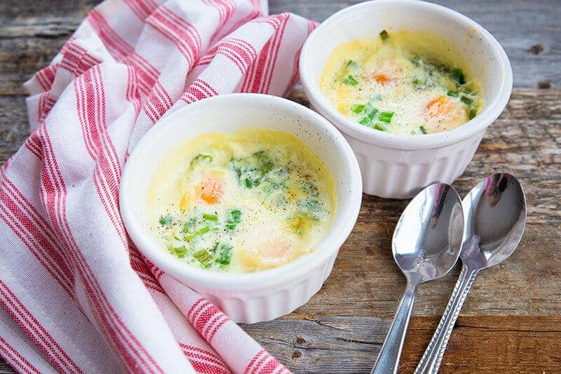 a kitchen cloth beside 2 soup bowl saucers with Shirred Eggs and spoons