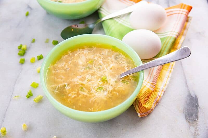 A bowl of Egg Drop Soup with spoon in it. Folded kitchen cloth underneath 2 fresh eggs