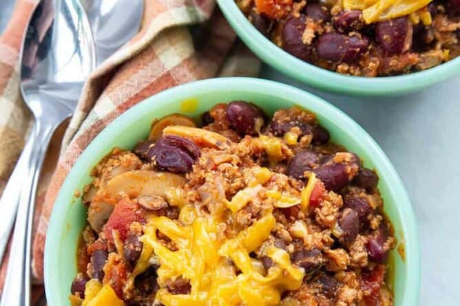 Ground Turkey Chili in small bowls with grated cheese on top, kitchen towel underneath 2 spoons