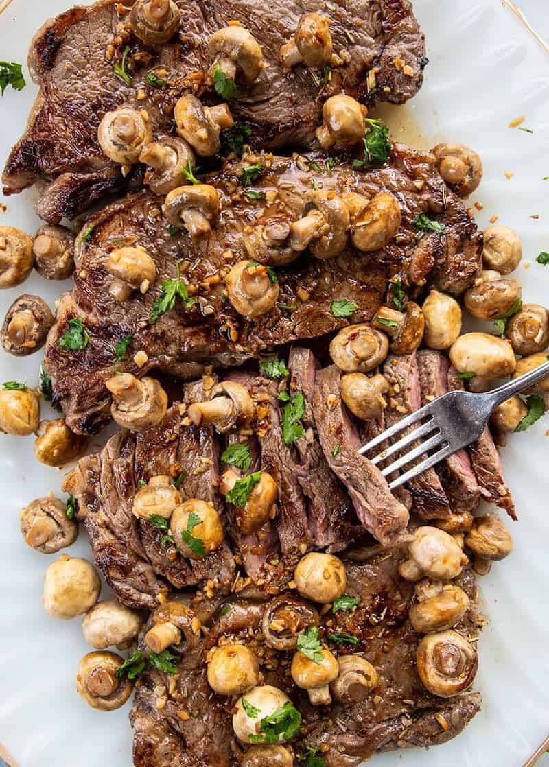 close up of fork holding slice of Cooked New York Strip Steak with Fried Garlic Mushrooms in a white plate