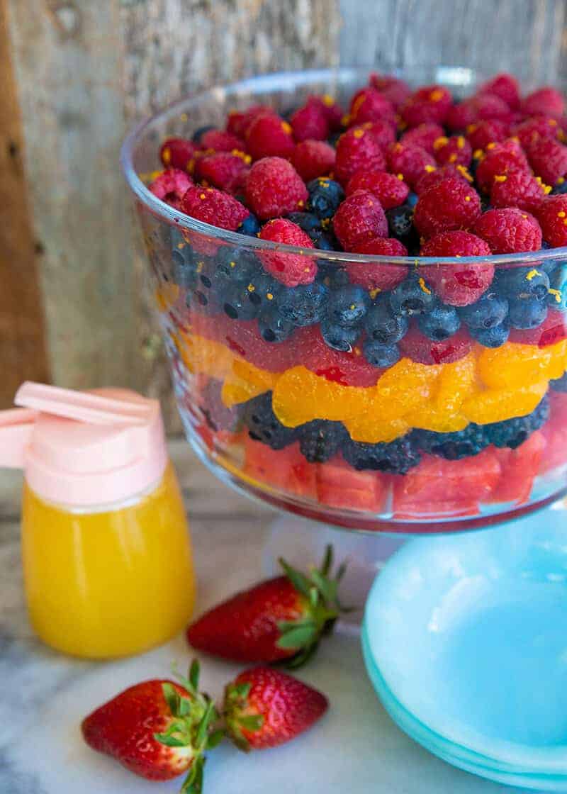 close up Summer Berry Fruit Salad in a trifle bowl. Blue dessert plates, fresh strawberries and container of orange juice dressing beside it.