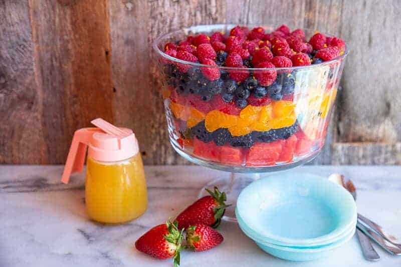 Summer Berry Fruit Salad in a trifle bowl. Spoons, blue dessert plates, fresh strawberries and container of orange juice dressing beside it.