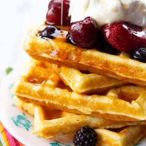 adding syrup to a stack of Homemade Belgian Waffles topped with some berries and Whipped Cream