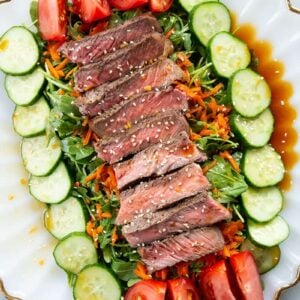 close up Asian Sesame Grilled Steak Salad in a white serving plate, drizzled with salad dressing and sprinkled with sesame seeds