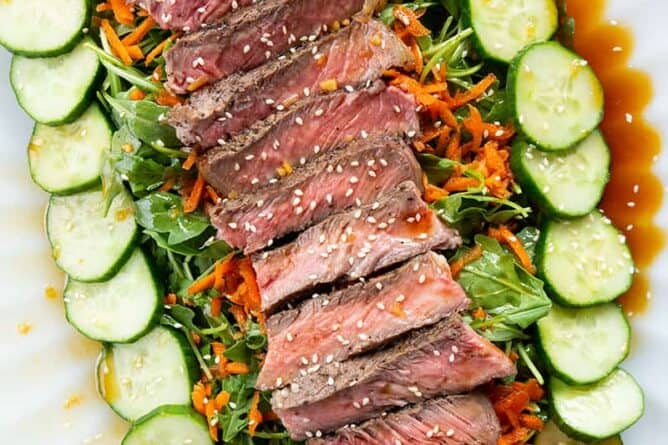 close up Asian Sesame Grilled Steak Salad in a white serving plate, drizzled with salad dressing and sprinkled with sesame seeds