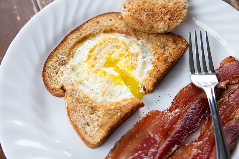 white plate with strips of bacon and Eggs in a Basket - crispy toasted bread with a gooey egg centre