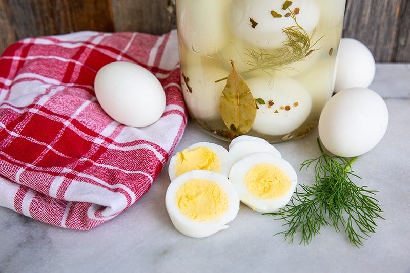 Pickled Eggs in sterilized 2-litre glass jar. Red checkered table cloth, boiled eggs and sprigs fresh dill around the jar