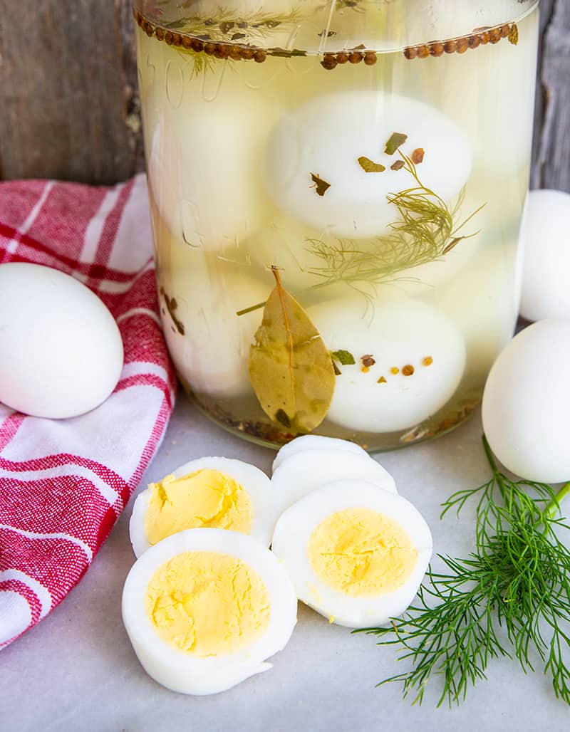 close up Pickled Eggs in sterilized 2-litre glass jar. Red checkered table cloth, boiled eggs and sprigs fresh dill around the jar