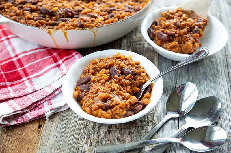 some serving of Cowboy Beans in white small plates, red checkered kitchen cloth and spoons on the side