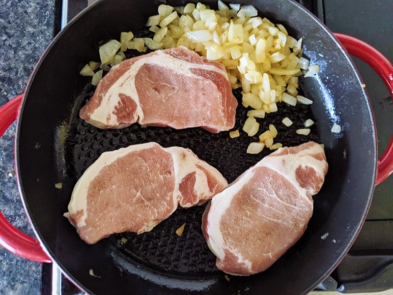 pork chops being cooked with onions in a red skillet