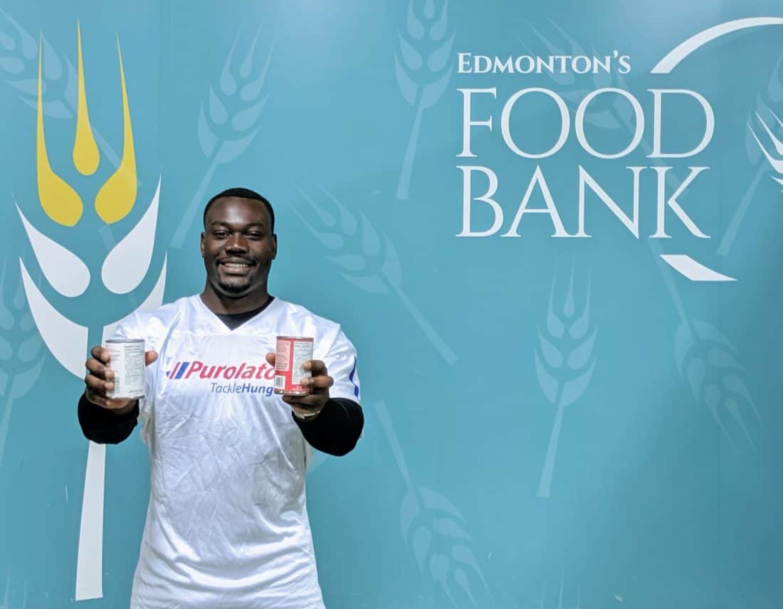 Kwaku holding 2 cans, Edmonton's Food Bank wallpaper on his background