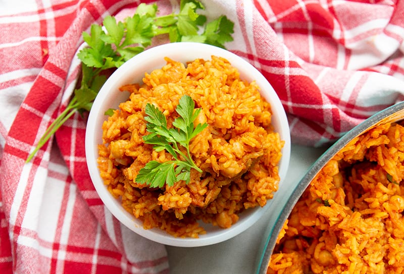 high angle shot red checkered tablecloth underneath a bowl of Jollof rice with chickpeas garnish with parsley