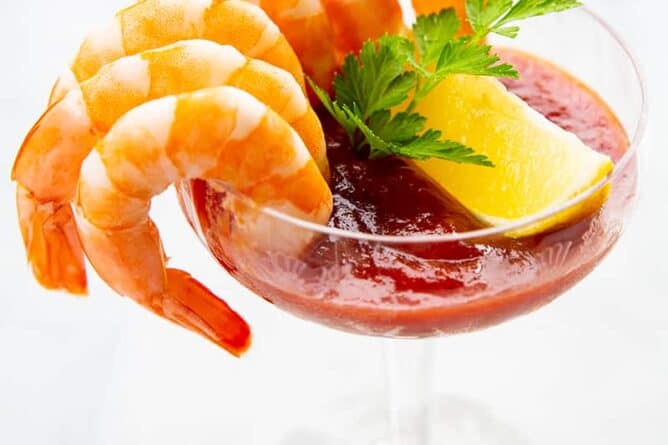6 cocktail shrimp around a serving glass with Cocktail Sauce garnish with a slice of lemon and parsley leaves