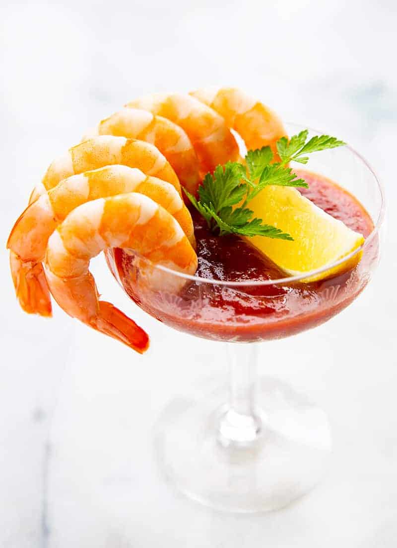 6 cocktail shrimp around a serving glass with Cocktail Sauce garnish with a slice of lemon and parsley leaves