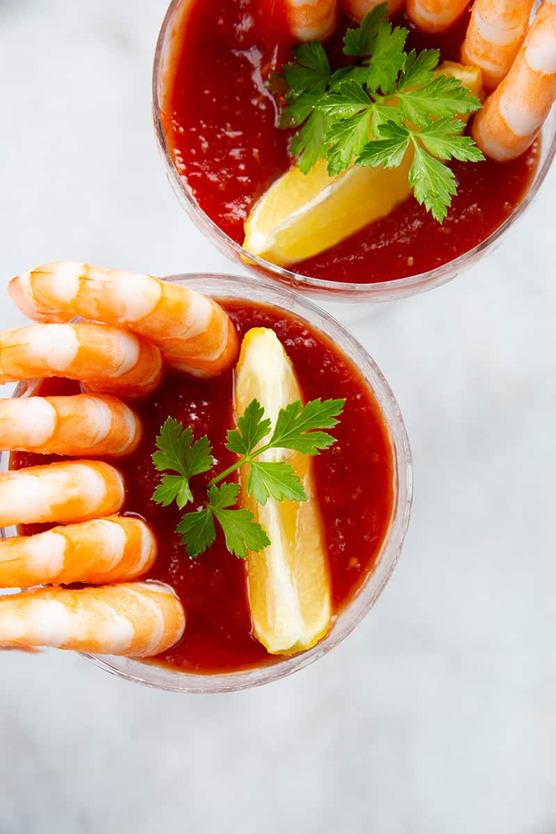 high angle shot of cocktail shrimp around a serving glass with Cocktail Sauce garnish with a slice of lemon and parsley leaves