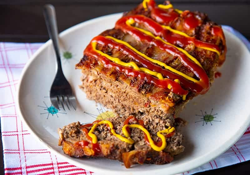 slice up Bacon Cheeseburger Meatloaf topped with cheese, mustard and ketchup in a white plate with fork