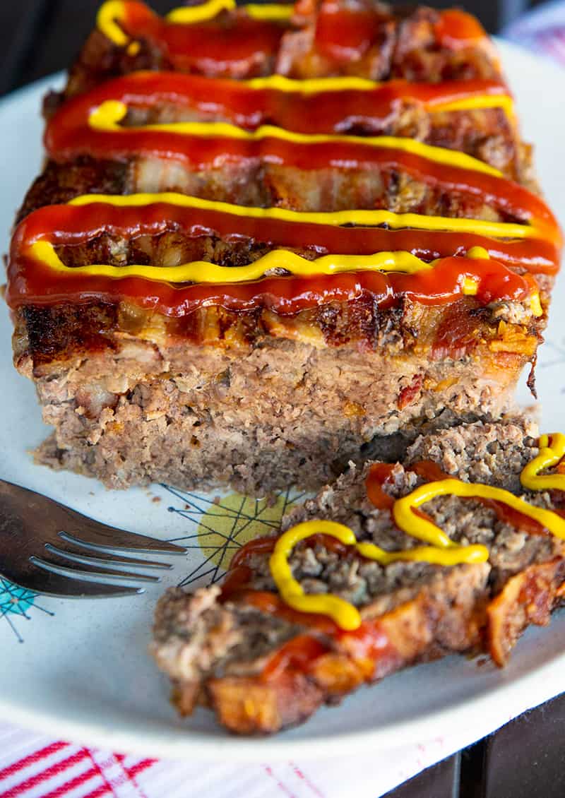 a plate of Bacon Cheeseburger Meatloaf slice up and topped with cheese, mustard and ketchup