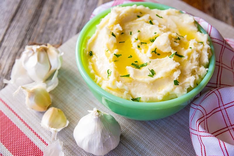 Buttery Garlic Mashed Potatoes in a mint green serving bowl, garlic head and cloves beside it