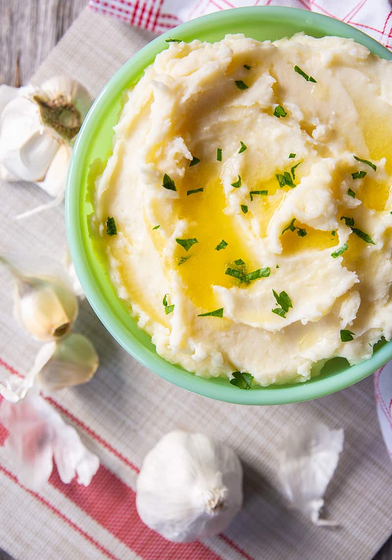 close up Buttery Garlic Mashed Potatoes in a mint green serving bowl, garlic head and cloves beside it