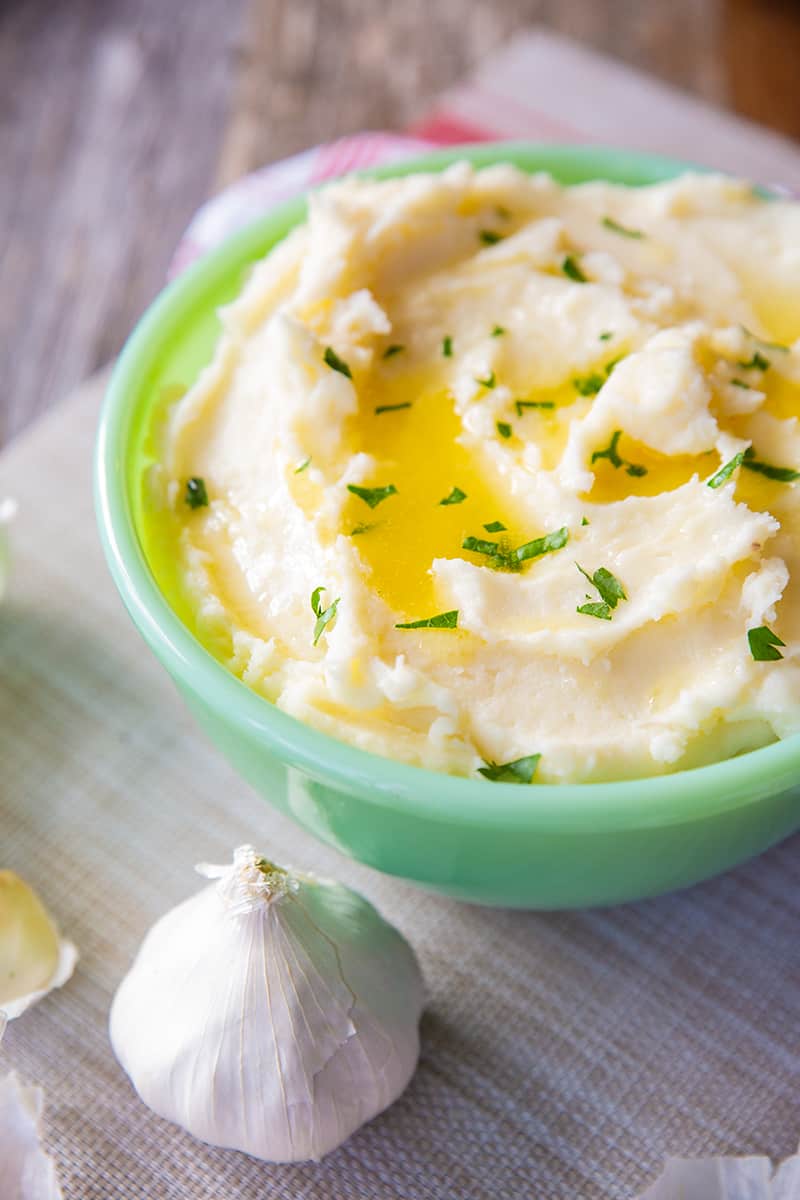 close up Buttery Garlic Mashed Potatoes in a mint green serving bowl and a garlic head beside it