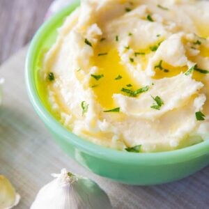 close up Buttery Garlic Mashed Potatoes in a mint green serving bowl and a garlic head beside it