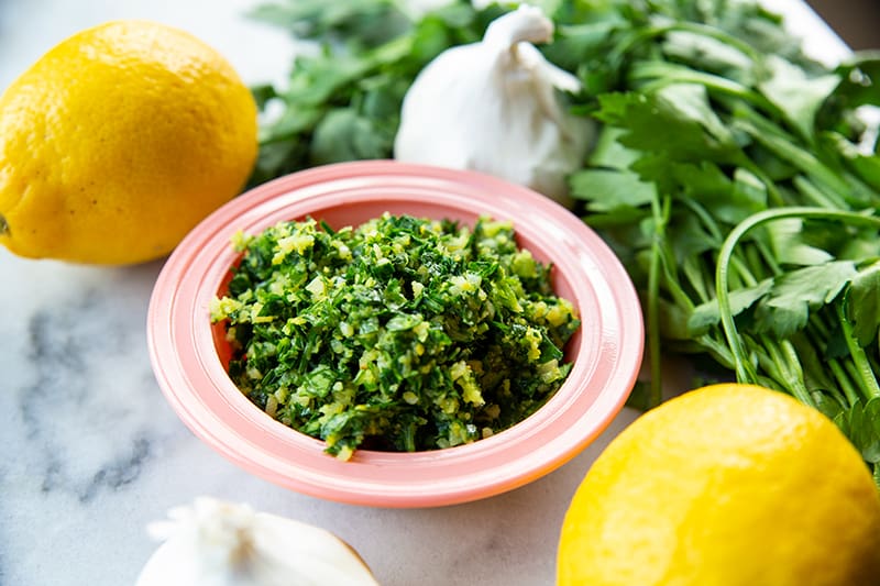 Classic Italian Gremolata in a pink saucer plate. 2 pieces of lemon, garlic head and parsley stems on background