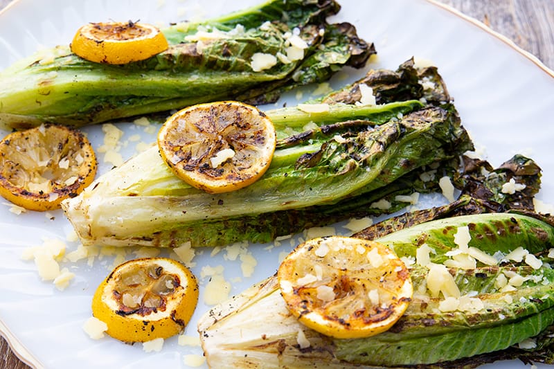 3 pieces of Parmesan Garlic Grilled Romaine in a white plate with some lemon slices on top and parmesan garlic toppings