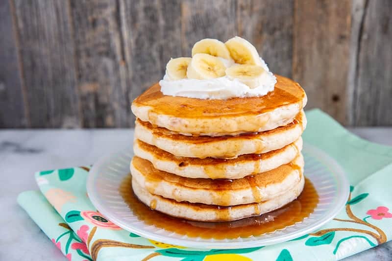 fluffy banana pancakes layer on a plate with maple syrup, whipped cream and sliced bananas on top