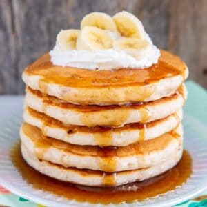 close up fluffy banana pancakes layer on a plate with maple syrup, whipped cream and sliced bananas on top