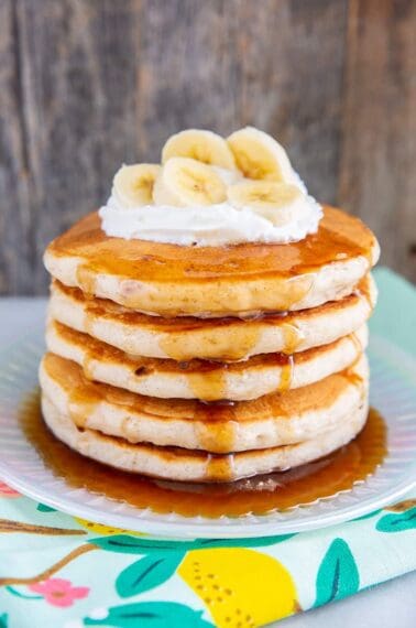 close up fluffy banana pancakes layer on a plate with maple syrup, whipped cream and sliced bananas on top