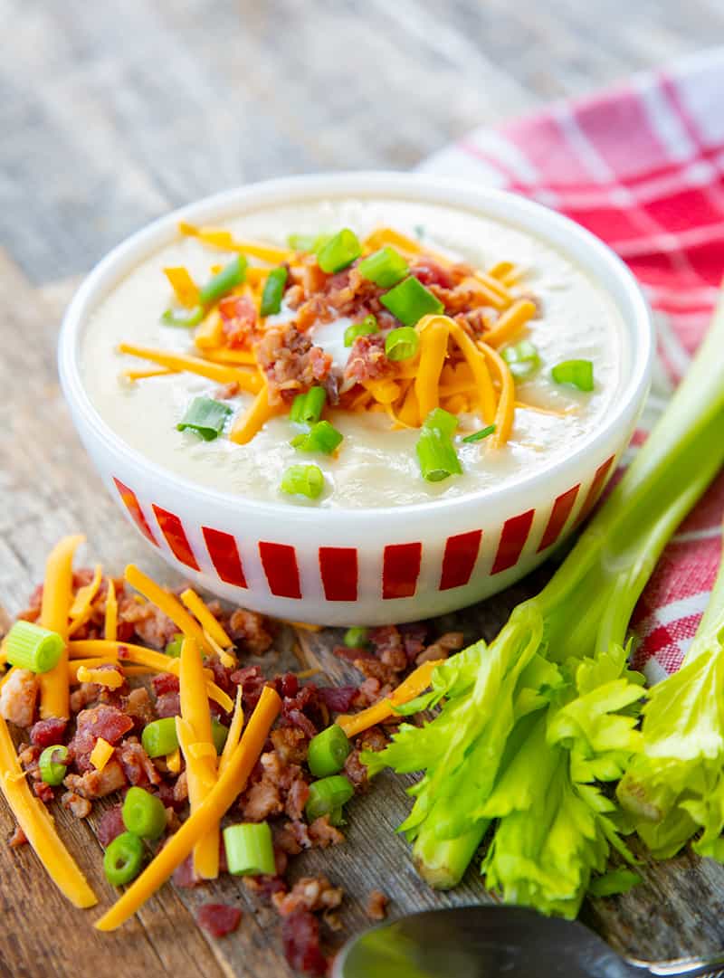 Super thick Cream of Celery Soup topped with shredded cheese, bacon bits and green onions in a white soup bowl. Raw celery stem and some toppings on its background