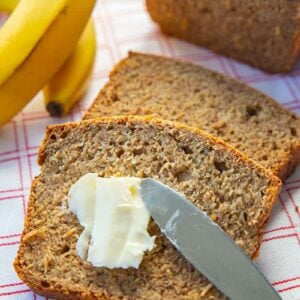 adding some spread into healthy banana bread slice using bread knife, ripe bananas and banana bread loaf on its background