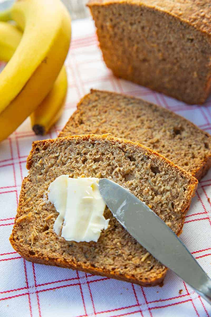 adding some spread into healthy banana bread slice using bread knife, ripe bananas and banana bread loaf on its background
