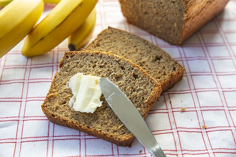 adding some spread into healthy banana bread slice using bread knife, ripe bananas and banana bread loaf on its background