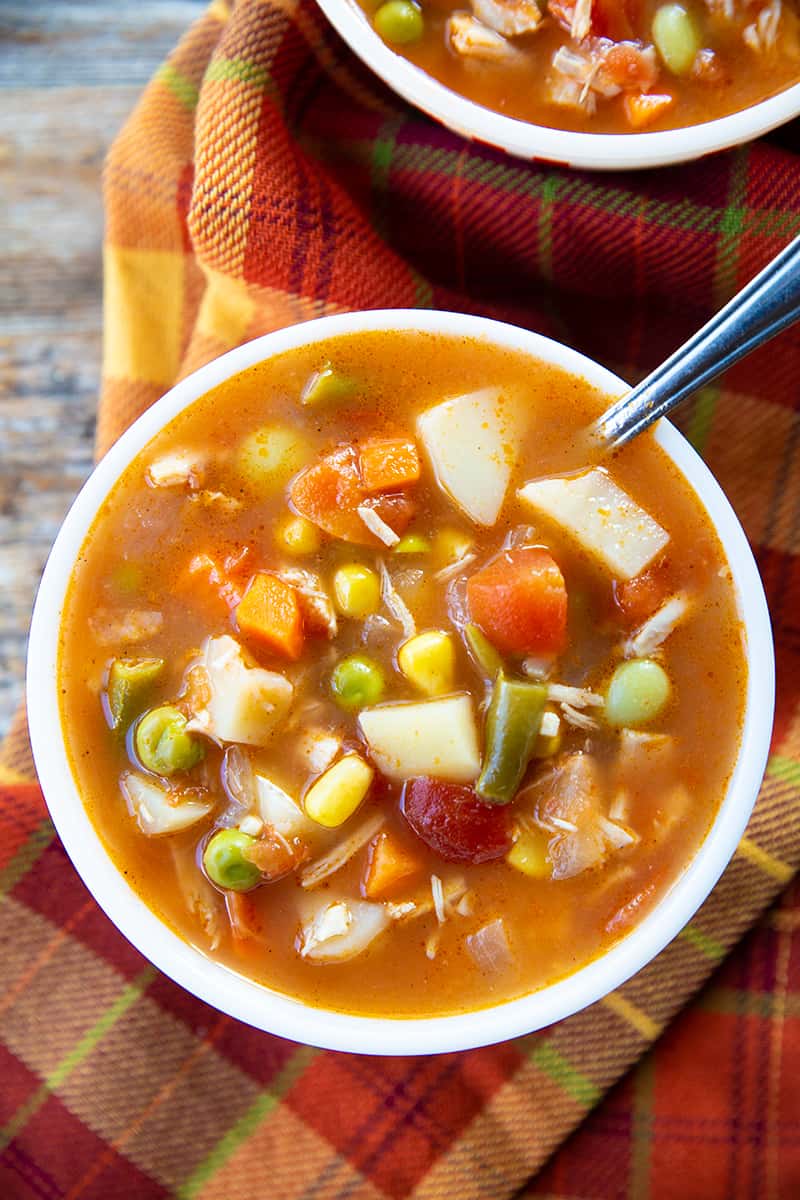 high angle shot of red orange kitchen cloth underneath a small serving bowl with Chicken Vegetable Soup and spoon on it