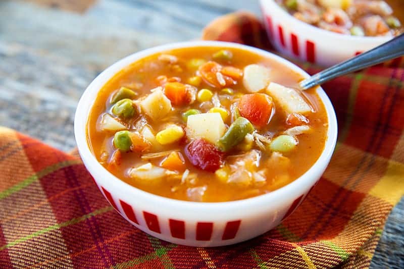 orange checkered tablecloth underneath a soup bowl filled with tomato based chicken vegetable soup with spoon