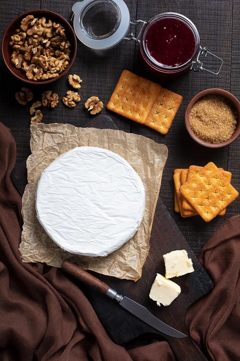 Walnut Raspberry Baked Brie ingredients on dark wood background with brown tablecloth on side