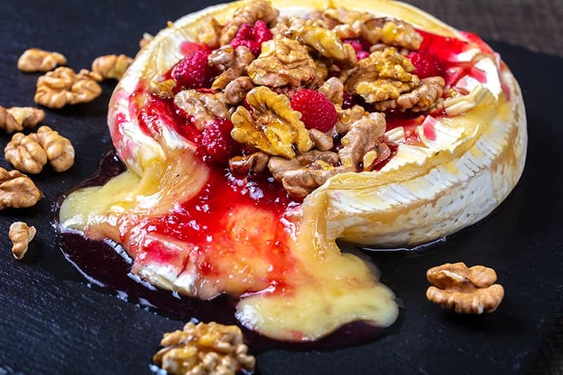 Walnut Raspberry Baked Brie in a black serving plate with raspberries and crunchy walnuts around