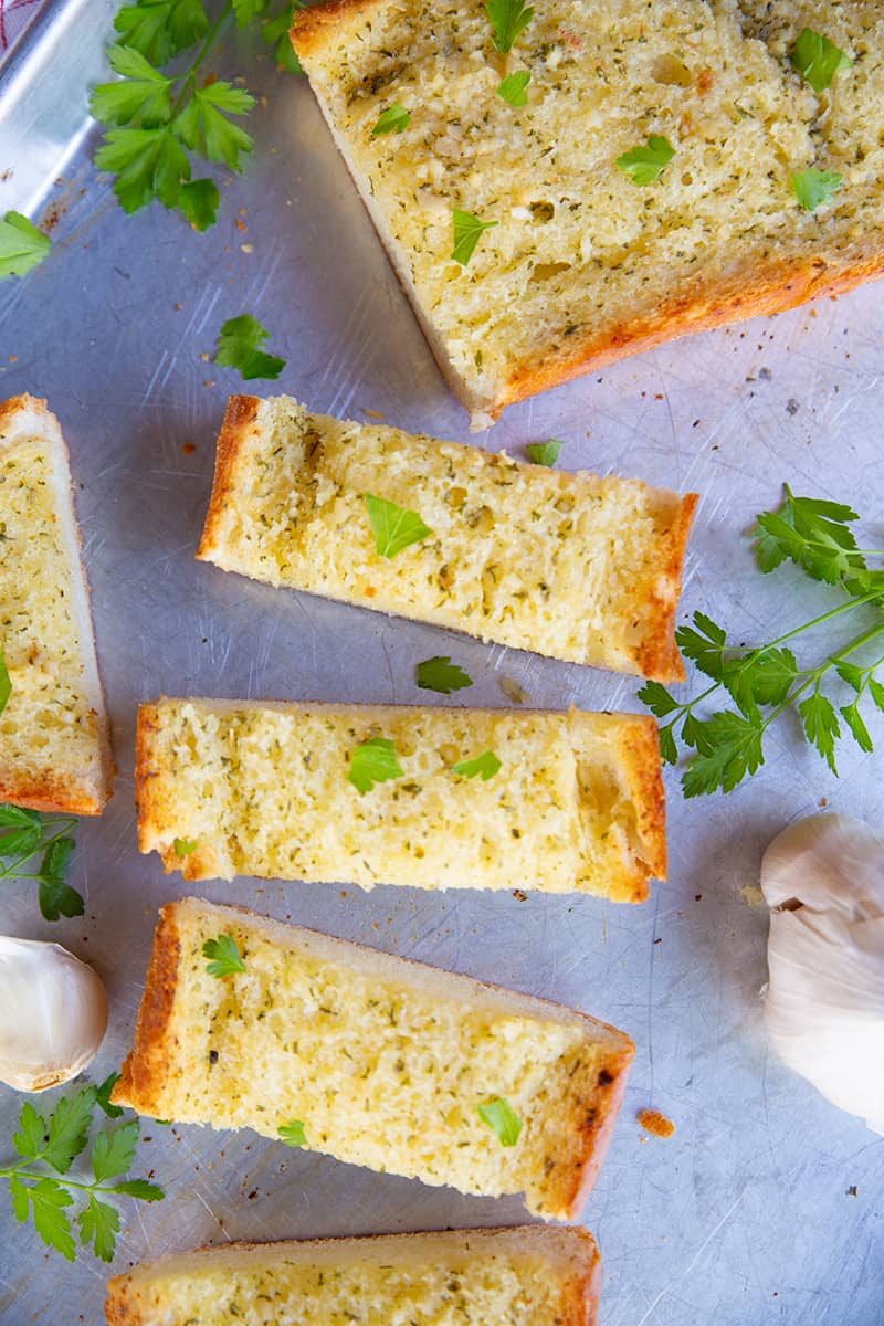 close up slices of Classic Homemade Garlic Bread in a baking sheet, pieces of garlic and some dried parsley leaves on its background
