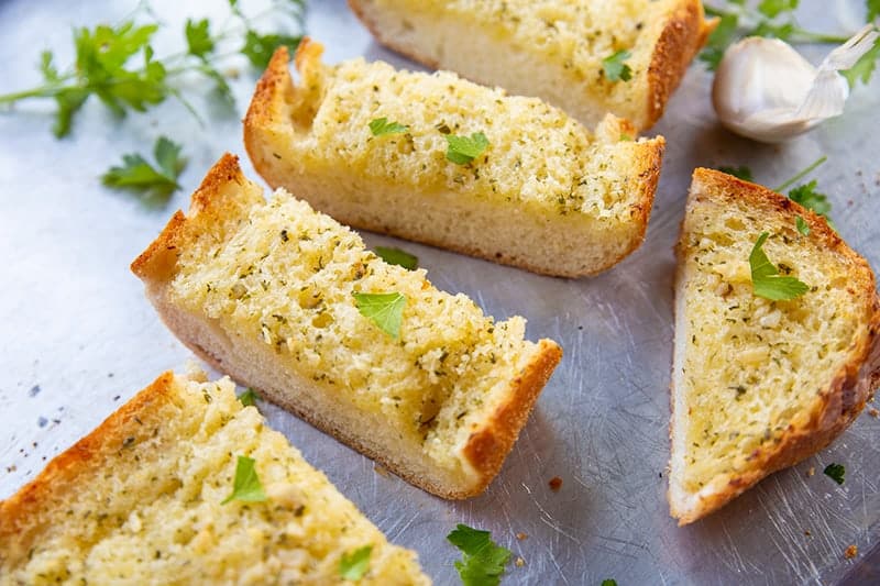 slices of Classic Homemade Garlic Bread in a baking sheet, a piece of garlic and some dried parsley leaves on its background