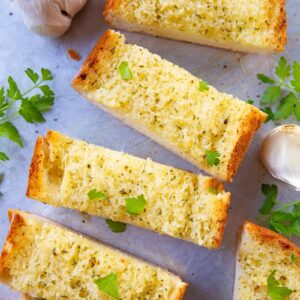 close up slices of Classic Homemade Garlic Bread in a baking sheet, pieces of garlic and some dried parsley leaves on its background