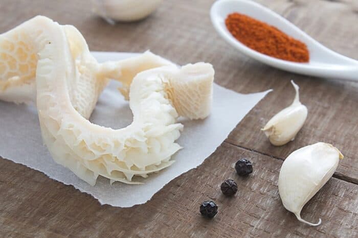 close up raw tripe on a parchment paper, black pepper corns, garlic cloves and powder seasoning on spoon beside it