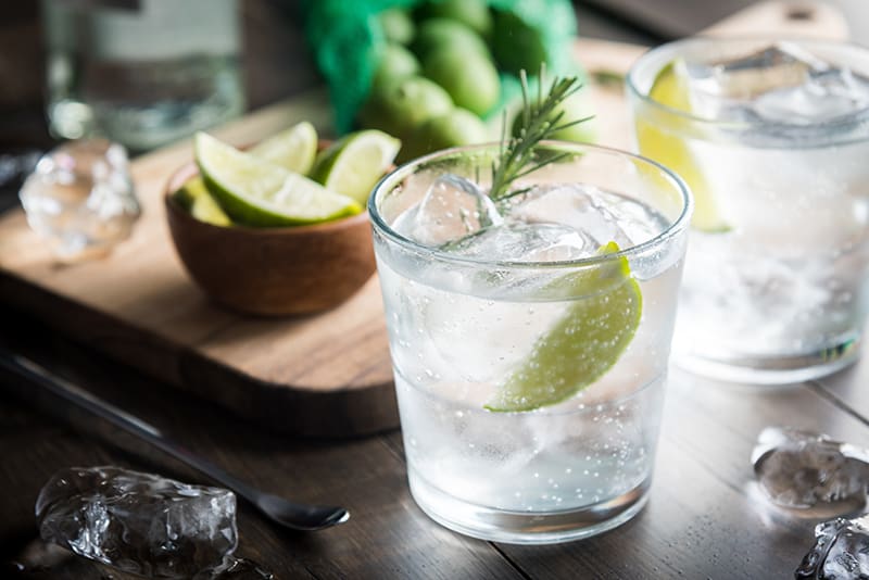 tonic water and lime on a clear glass with some ice cubes, some slices of lime in a wooden cup on the background