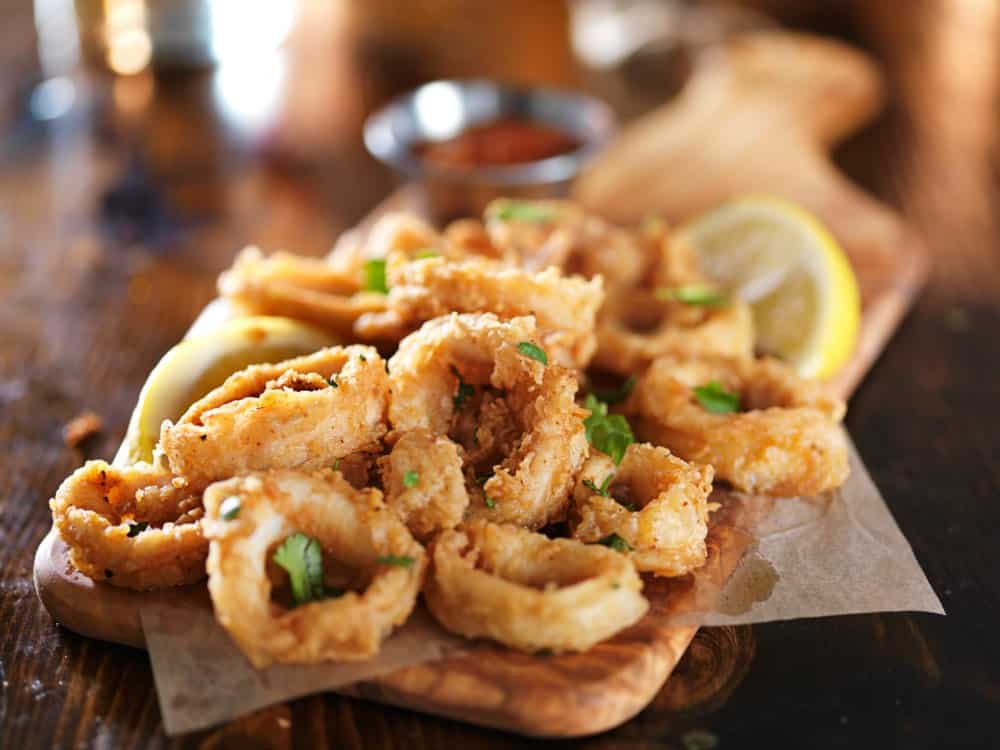 wooden cutting board underneath a layer of kitchen towel with breaded fried Calamari, a cup of marinara sauce on its background