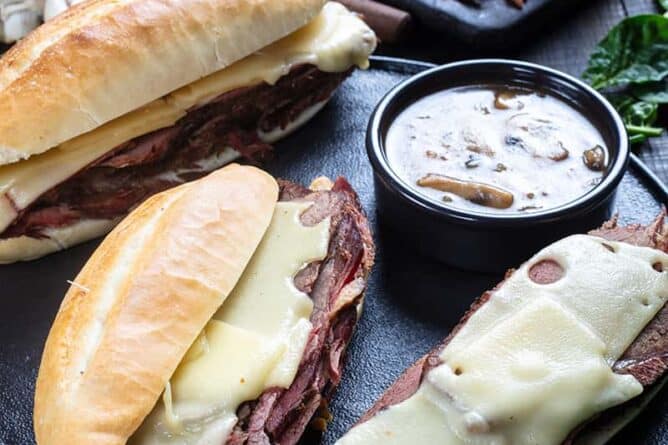 close up Cheesy Mushroom French Dip Sandwiches and a cup of gravy on a large serving plate