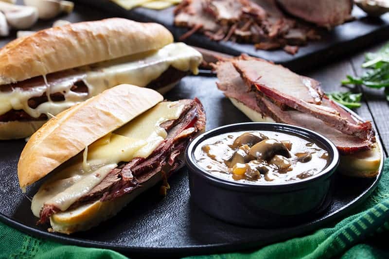 close up Cheesy Mushroom French Dip Sandwiches and a cup of gravy on a large serving plate